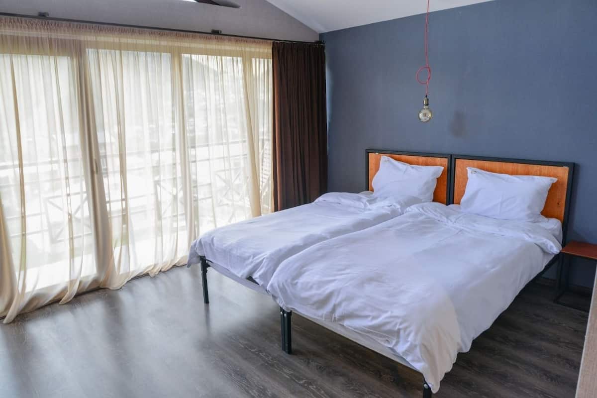 A double bed with white linens in a room with a wooden floor and a view through sheer curtains.