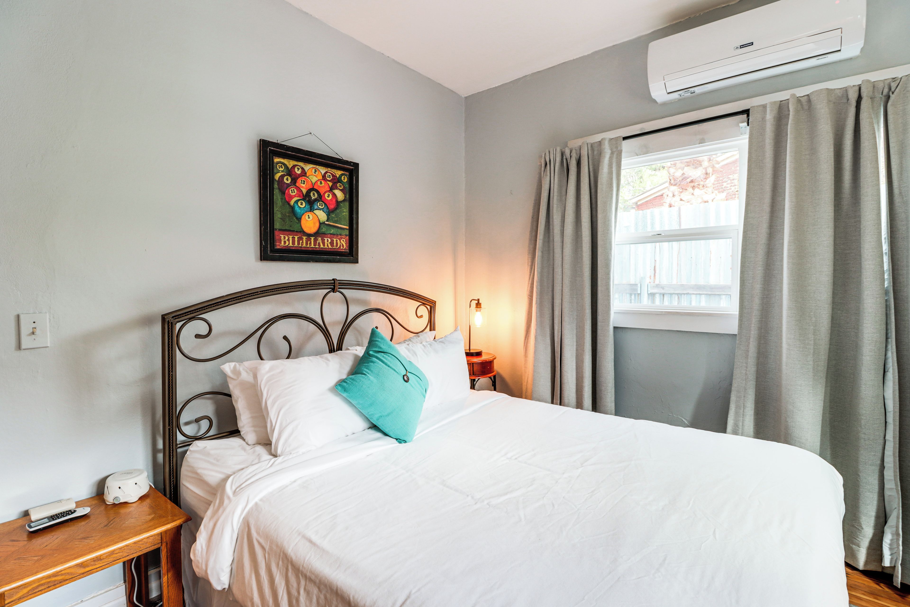 A neatly made bed with white bedding and a teal pillow, a framed billiards poster on the wall, a bedside table with a lamp, and a window with curtains.