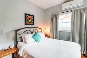 Een netjes opgemaakt bed met wit beddengoed en een turquoise kussen, een ingelijst poster van een biljartspel op de muur, een nachtkastje met een lamp en een raam met gordijnen.