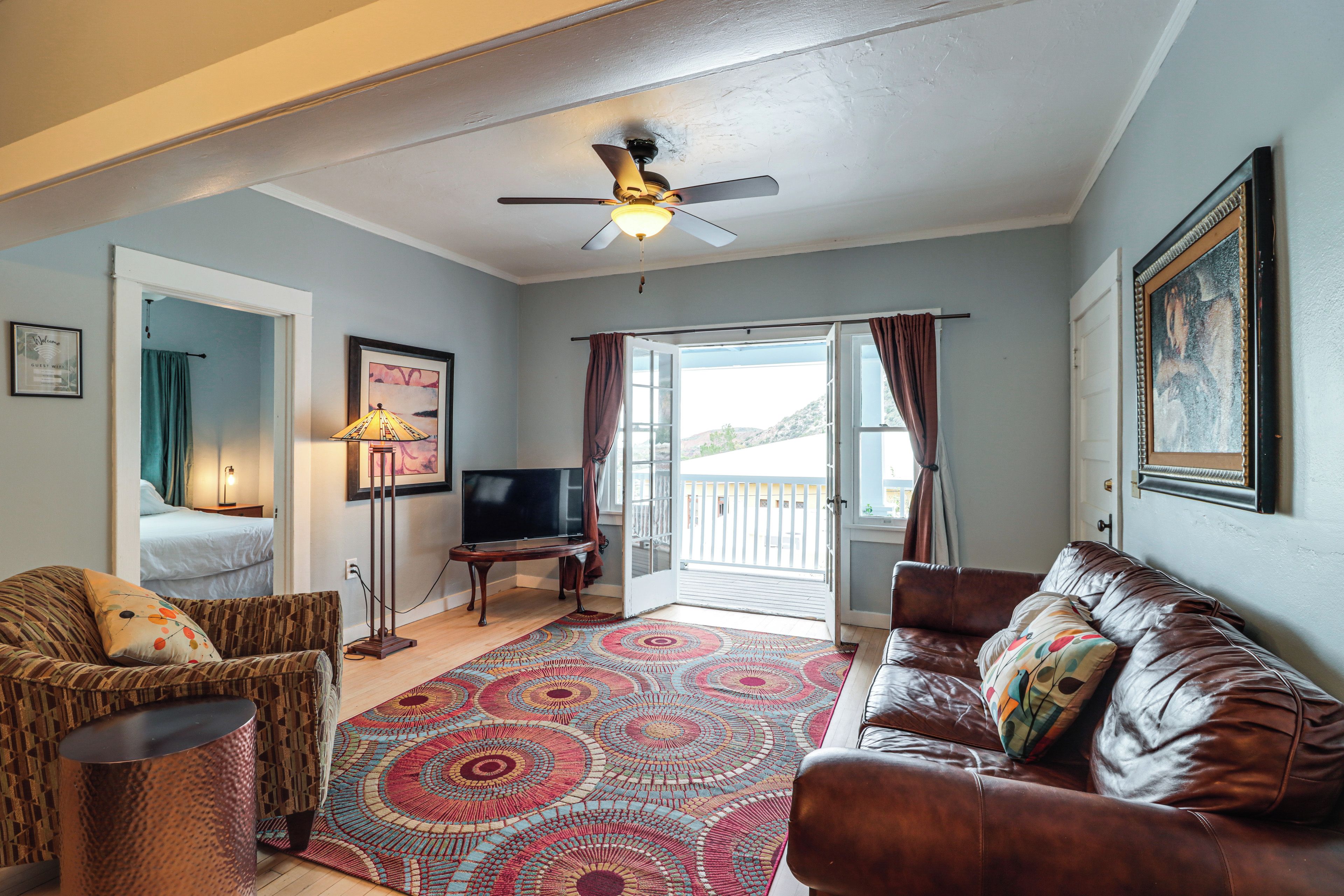 A living room with a ceiling fan, a flat-screen TV, a patterned rug, a leather couch, and a bed in the adjacent room.