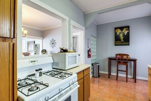 Een keuken met een witte kookplaat, een magnetron en een spoelbak. Er staat een stoel en een tafel met een poster aan de muur.