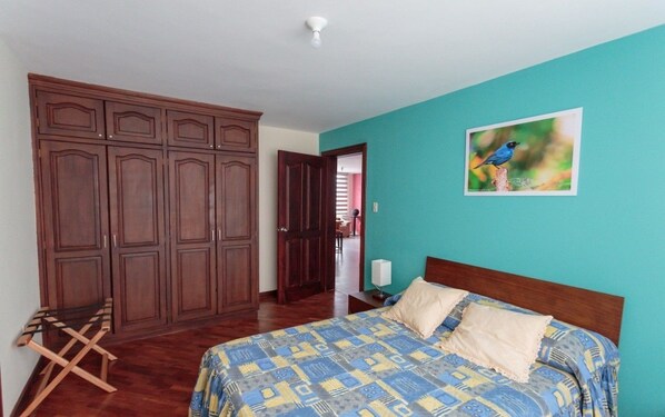 A bedroom with a bed, a wooden wardrobe, and a painting of a bird on the wall. - Apartments Cumbaya Quito Ec (Cumbayá)