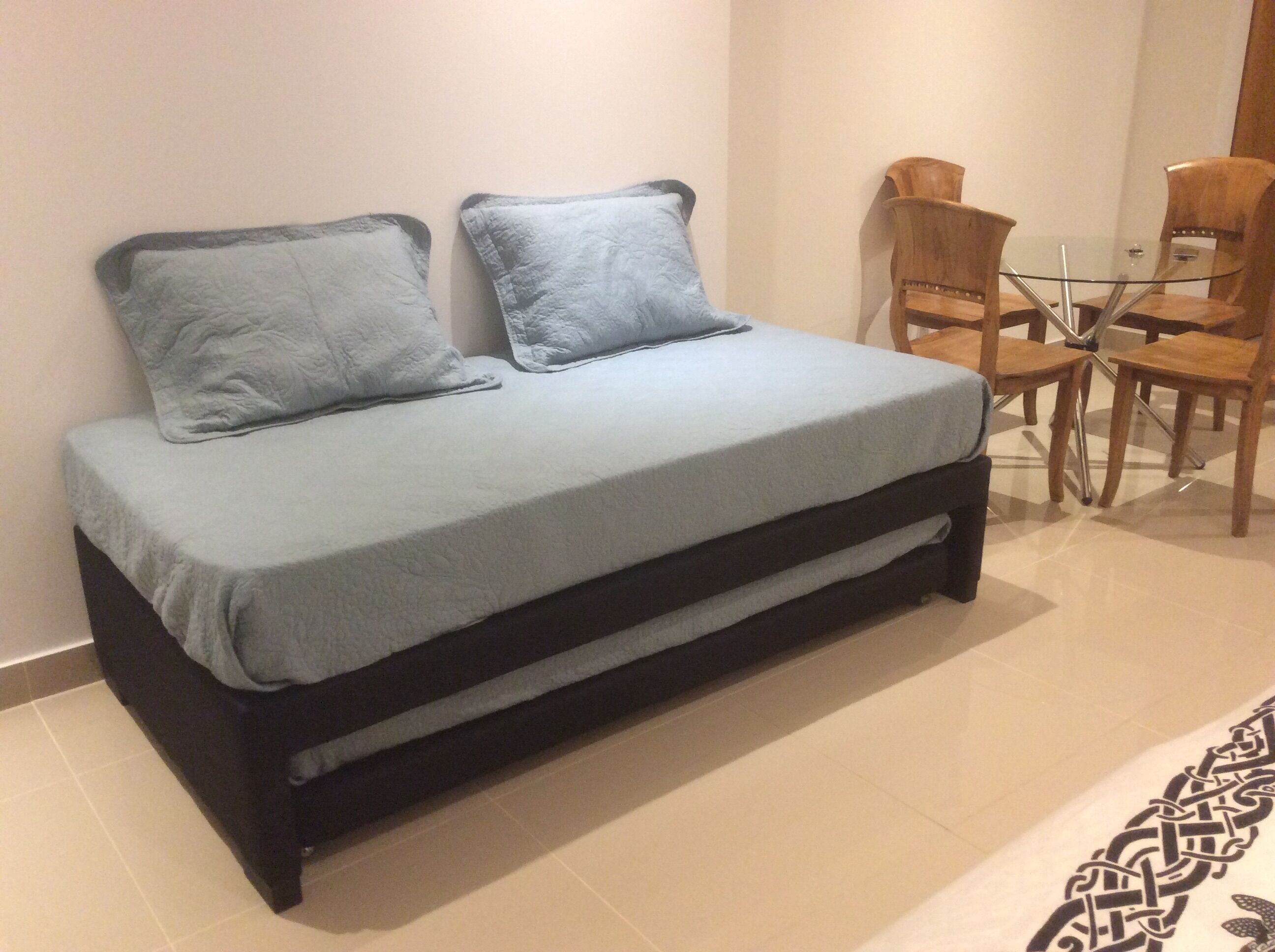 A bed with a grey mattress and two pillows, a wooden table with chairs, and a patterned rug.