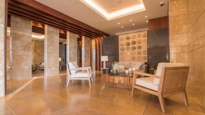 A spacious lobby with a modern design, featuring a seating area with two white chairs and a wooden side table, a decorative wall with circular patterns, and a large mirror reflecting the space. - 1703 Apartamento Luxury en Edificio H2 Condominio (Cartagena)