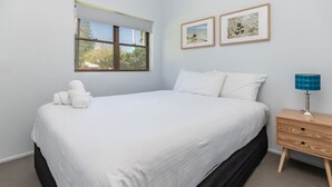 A neatly made bed with white linens, a wooden nightstand with a lamp, and two framed pictures on the wall. - Two Storey Apartment Sandcastle 7 (Tuncurry)