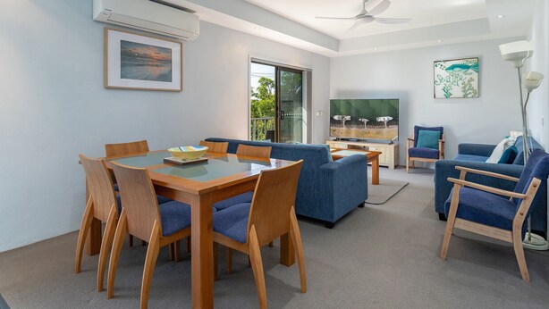 A modern living room with a glass dining table, blue sofas, and a flat-screen TV. - Two Storey Apartment Sandcastle 7 (Tuncurry)