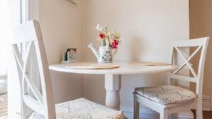 Una mesa redonda blanca con dos sillas blancas, un florero con flores y un cuadro en la pared.