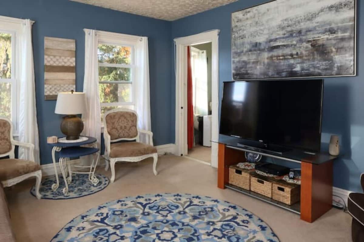 A living room with a large flat-screen TV on a wooden stand, two armchairs, a small side table with a lamp, and a patterned rug.
