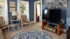 Un salón con un televisor de pantalla plana grande sobre un soporte de madera, dos sillones, una mesita auxiliar con una lámpara y una alfombra estampada.