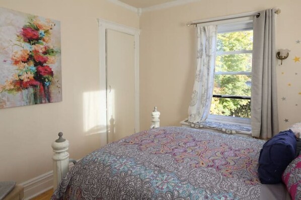 A bedroom with a patterned bedspread, a window with curtains, a white door, and a wall-mounted star decoration. - Montclair Apartment (Montclair)
