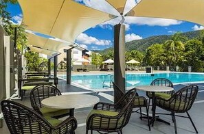 Relax in a shaded outdoor seating area with a round table and wicker chairs, overlooking a swimming pool surrounded by lush greenery and hills.