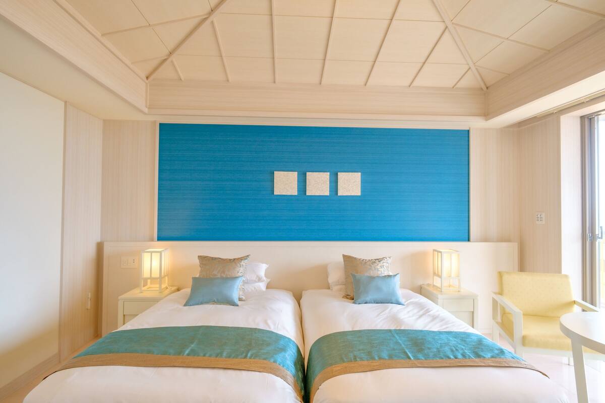 Ein Hotelzimmer mit zwei Betten, einer blauen Akzentwand, Nachttischlampen, einem gelben Sessel und einem kleinen Tisch.