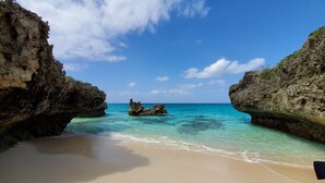 Une plage à l’eau turquoise et limpide, bordée d’un rivage sablonneux et ponctuée de formations rocheuses.
