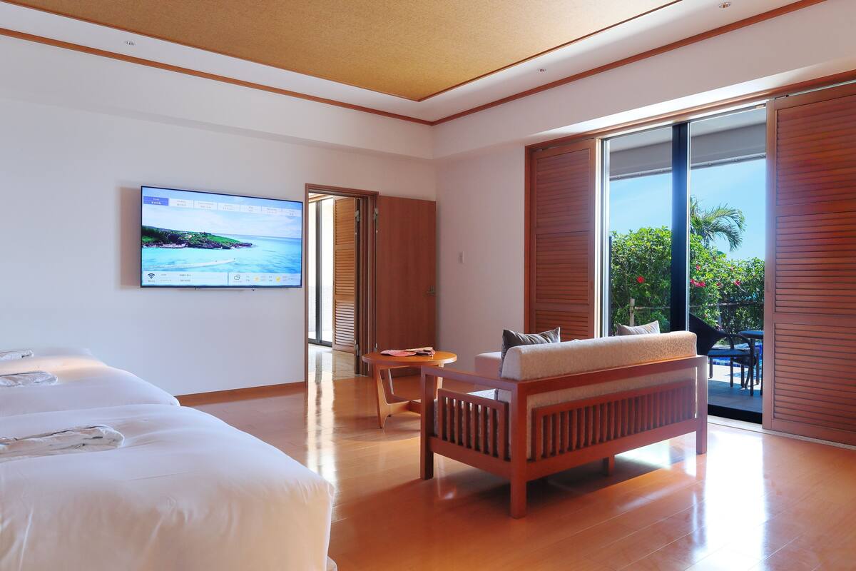 Ein modernes Hotelzimmer mit einem großen Bett, einer hölzernen Sitzbank, einem Fernseher und einer Schiebeglastür zum Außenbereich.
