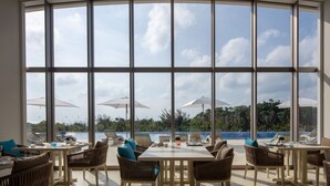 Une salle à manger baignée de lumière naturelle, dotée de grandes fenêtres, de sièges en plein air et de parasols, offrant une vue sur une piscine et un paysage tropical.