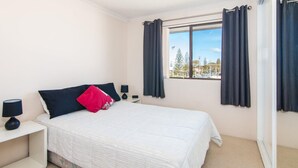A bedroom with a bed, two bedside tables, a window with curtains, and a view of trees and buildings outside. - Seabreeze 4 - Opposite Tuncurry Bowling Club! (Tuncurry)