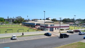 A parking lot with several cars, a building with a flat roof, and a grassy area in the background. - Seabreeze 4 - Opposite Tuncurry Bowling Club! (Tuncurry)