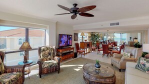 A spacious living room with a television, comfortable seating, a dining area, and a ceiling fan. - Upgraded Beachfront Condo Close to Shops, and Restaurants - Pool, Hot Tub, Beach, and Private Balcony (Madeira Beach)