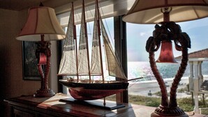 A model ship on a table with two lamps, a framed picture, and a view of the ocean through a window. - Upgraded Beachfront Condo Close to Shops, and Restaurants - Pool, Hot Tub, Beach, and Private Balcony (Madeira Beach)