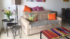 Een woonkamer met een beige bank, kleurrijke kussens, een vaas met bloemen, een lamp en een kleurrijk bijzettafeltje.