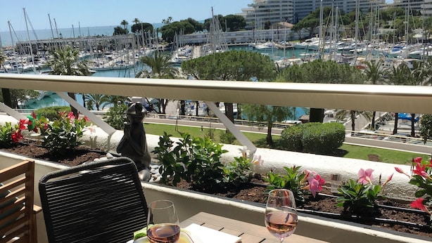 Een balkon met een tafel, twee borden eten en wijnkaraffen, met uitzicht op een jachthaven met boten en gebouwen.