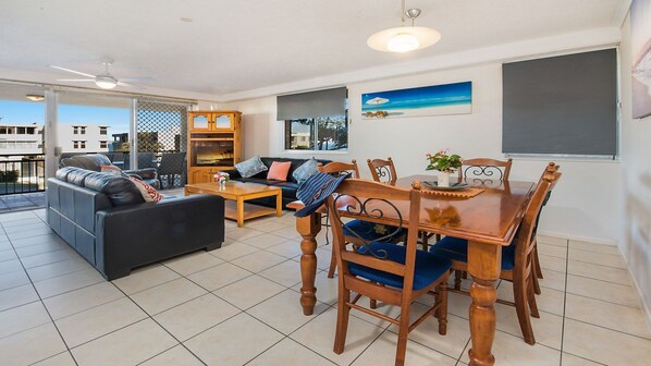 A spacious living room with a sofa, a dining table with chairs, and a TV mounted on the wall.