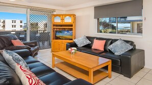 A living room with a sofa, armchairs, a coffee table, and a TV on a wooden cabinet. - Kokomo Unit 1 Kings Beach QLD (CALOUNDRA)