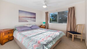A bedroom with a bed, a nightstand, a chair, a window with curtains, and a ceiling fan. - Kokomo Unit 1 Kings Beach QLD (CALOUNDRA)