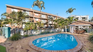 A pool area with a brick border, surrounded by a fence and palm trees, with a building in the background. - Kokomo Unit 1 Kings Beach QLD (CALOUNDRA)