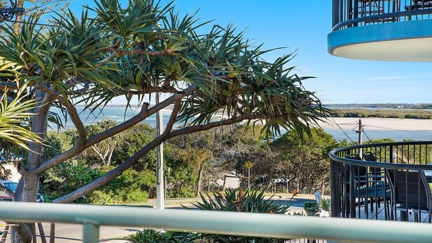 A balcony with a view of a beach and ocean, surrounded by tropical plants and a modern building structure. - Kokomo Unit 1 Kings Beach QLD (CALOUNDRA)