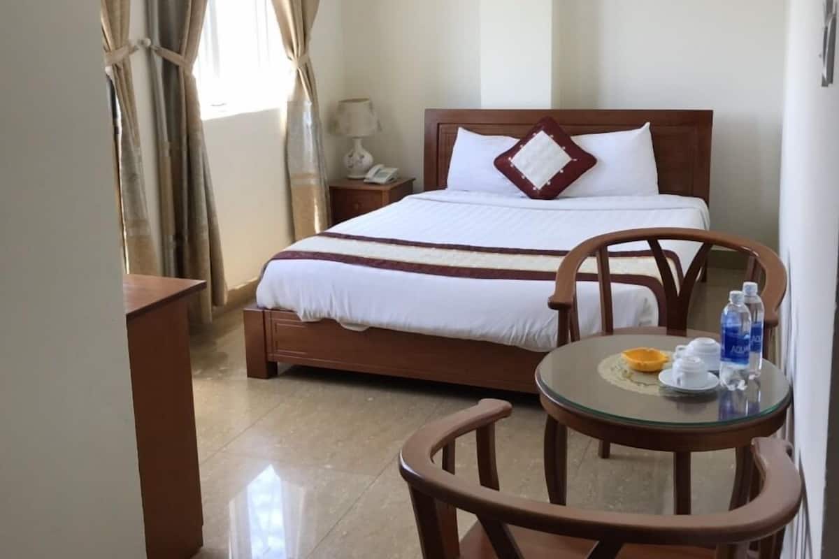 A hotel room with a bed, a desk, a chair, and a small table with a bottle and a cup.