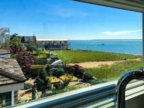 A coastal view from a window overlooking a sandy beach, a grassy area, and a few buildings. - Beachfront Studio with Spectacular Water Views (Provincetown)