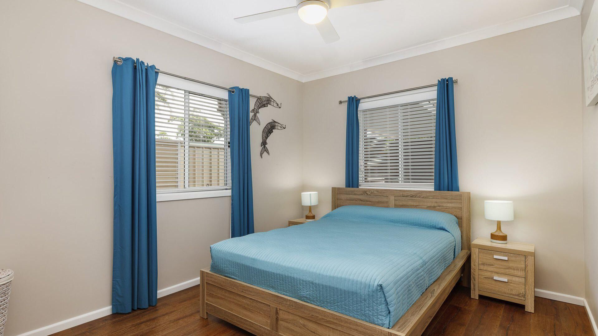 A bedroom with a wooden bed, two bedside tables with lamps, a ceiling fan, and a window with blue curtains.