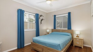 A bedroom with a wooden bed, two bedside tables with lamps, a ceiling fan, and a window with blue curtains. - Sandy Toes - park the car and walk everywhere! (Forster)
