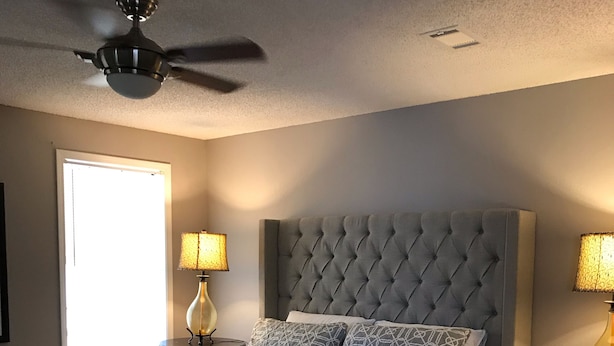Un dormitorio con cabecera capitonada, una cama grande con ropa de cama estampada, dos lámparas de noche, un ventilador de techo y una ventana con luz natural.