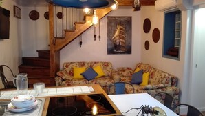 A cozy living room with a wooden staircase, a floral sofa, a dining table with a stove, and a wall-mounted air conditioner.