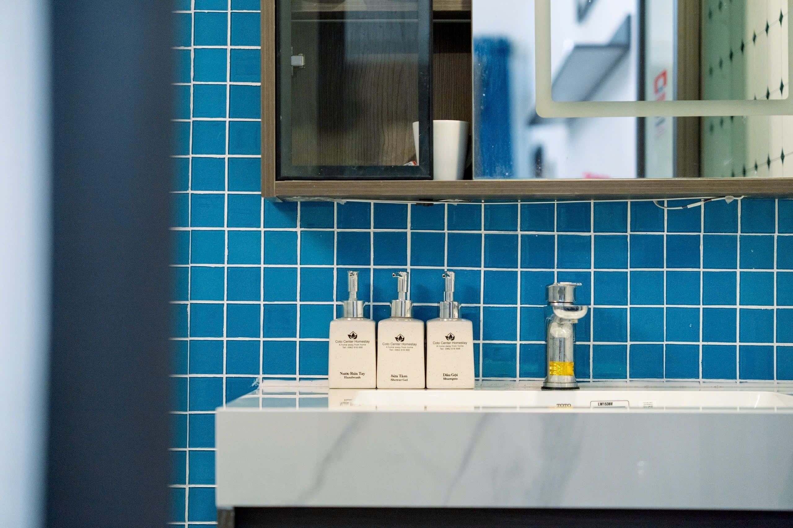 Une salle de bain avec un mur recouvert de carreaux bleus, un miroir et trois flacons de produits de toilette posés sur le lavabo.