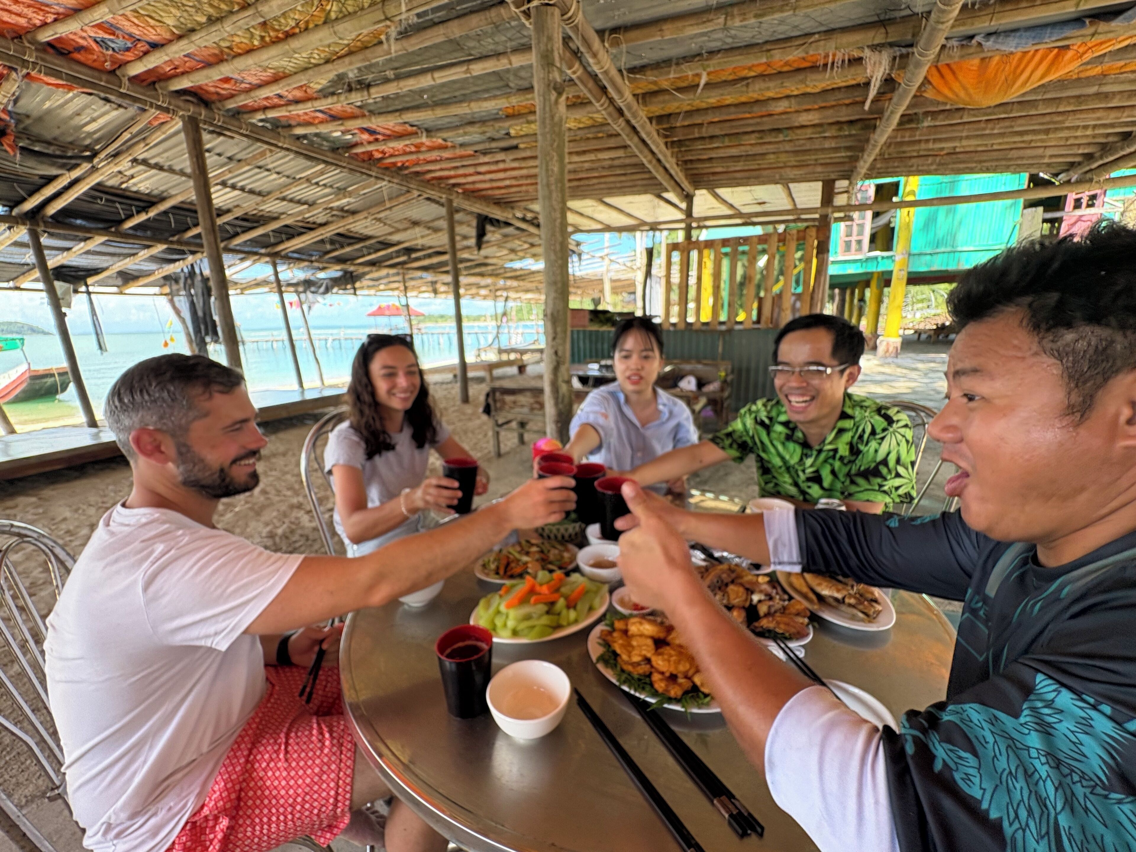 Un groupe de personnes est assis autour d’une table ronde, où sont disposés de la nourriture et des boissons. Elles se trouvent sous une zone abritée, d’où l’on peut admirer la mer.