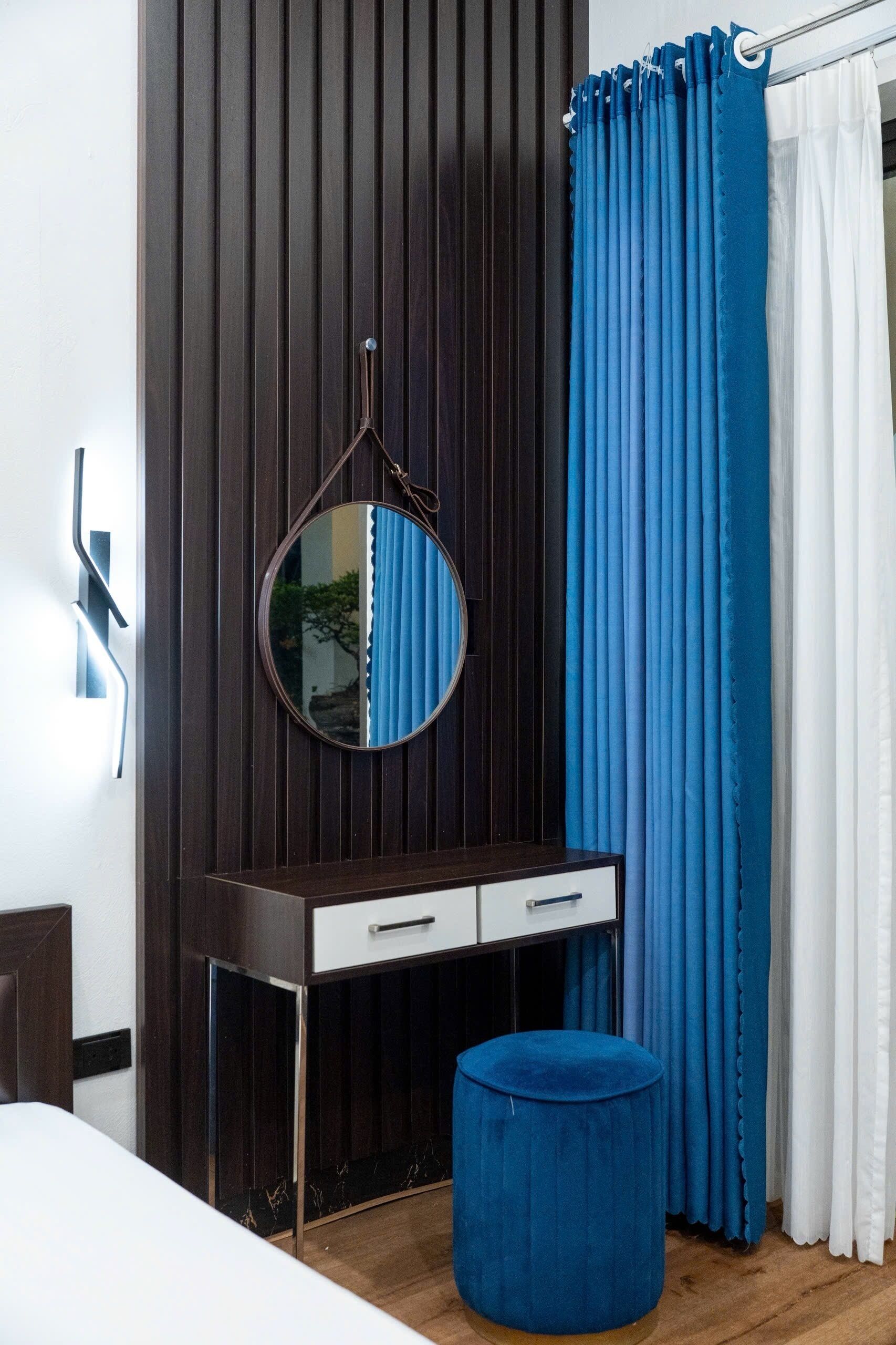 A bedroom with a dark wooden wardrobe, a round mirror, a white bedside table with drawers, a blue ottoman, and a window with blue and white curtains.