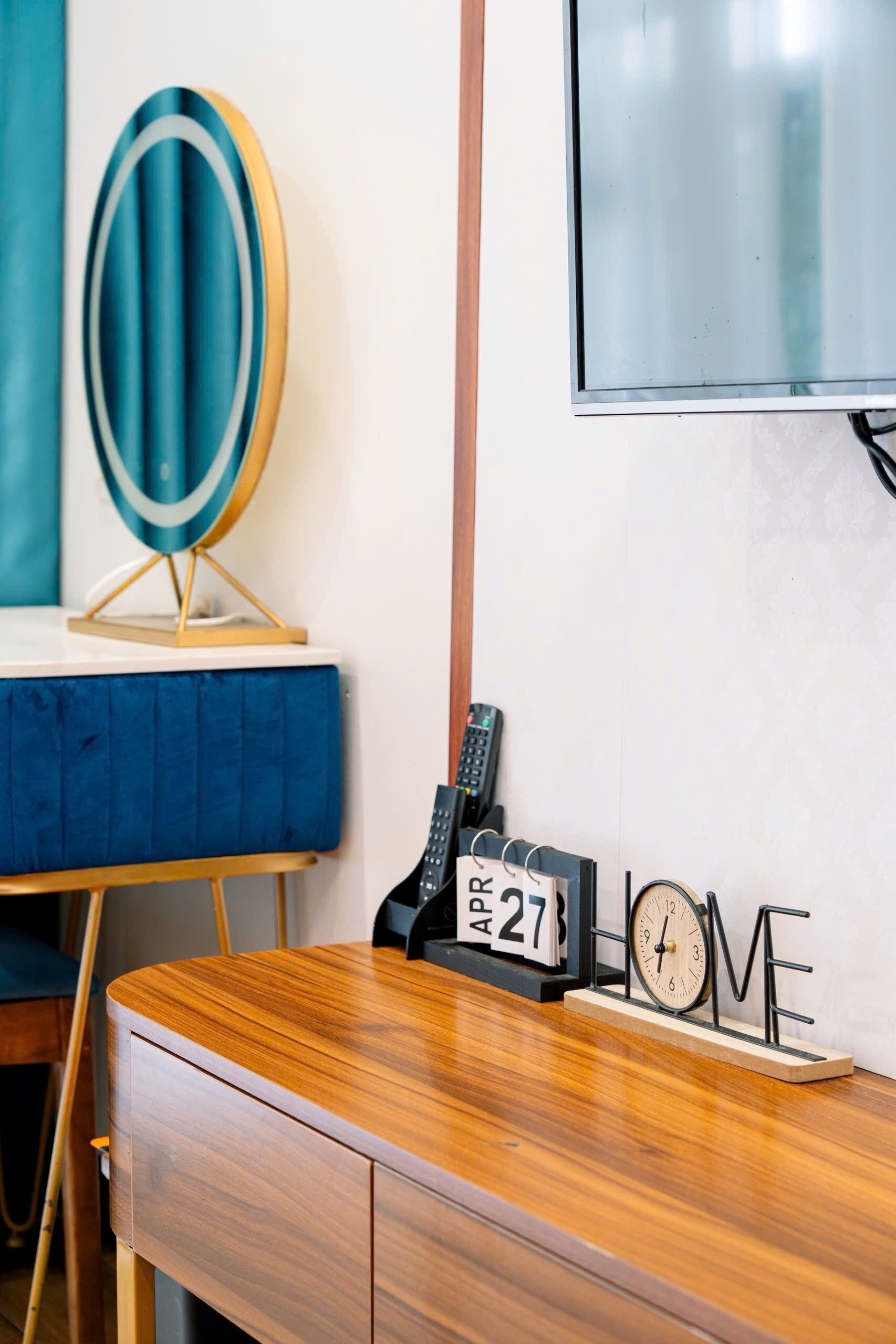 A room with a wooden desk, a blue chair, a wall-mounted TV, a clock, and a calendar.