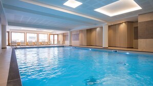 An indoor swimming pool with clear blue water, a seating area with chairs, and a modern ceiling design.