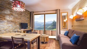 A modern room with a wooden dining table, chairs, a sofa, and a large window offering a mountain view.