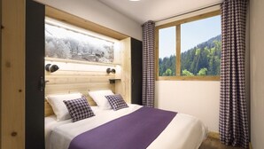 A bedroom with a wooden headboard, a bed with white and purple bedding, a window with a view of a forest, and patterned curtains.