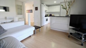 A modern living room with a sofa, a small table, a TV on a cart, and a kitchen area with cabinets and a microwave.