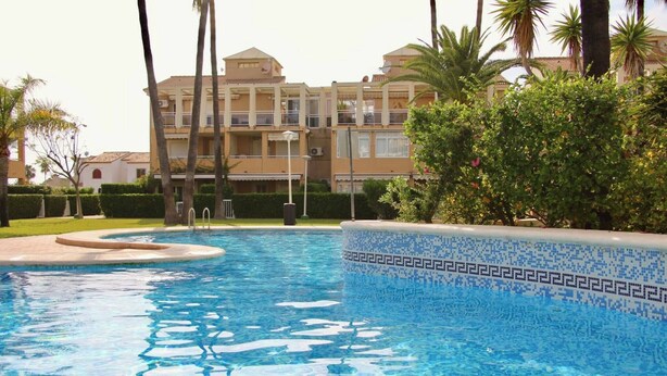 A swimming pool with a tiled edge, surrounded by greenery and a building with balconies in the background.