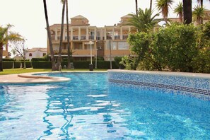 Une piscine dont le bord est revêtu de carreaux, entourée de verdure, avec un bâtiment doté de balcons en arrière-plan.