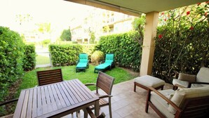 An outdoor patio with a wooden table and chairs, a cushioned sofa, and a view of a well-maintained garden and neighboring buildings.