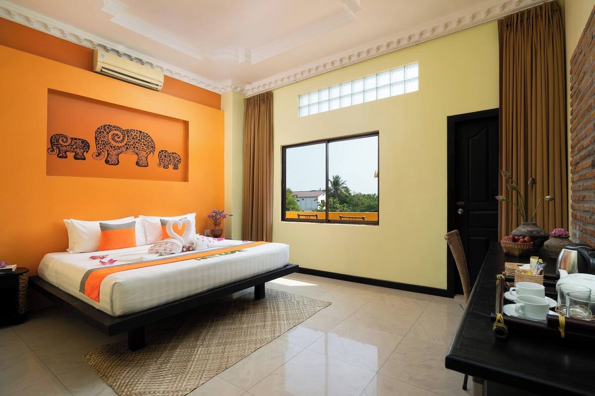 A hotel room with a bed, a desk with a chair, a window with a view, and a wall with a decorative elephant mural.