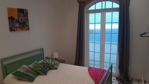 Un dormitorio con cama, ventana con cortinas, un cuadro en la pared y vista al mar.