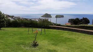 Un área verde con un columpio y un muro de piedra, con vistas a un paisaje costero con casas y cielo despejado.